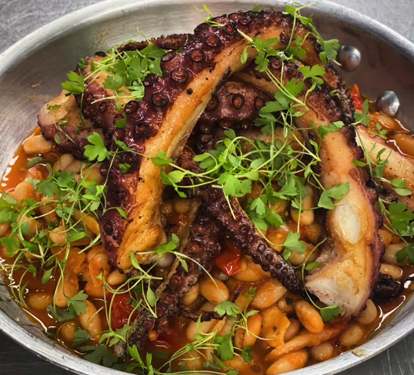 Screen Shot 2022-04-25 at 4.54.58 PM A dish with pieces of grilled octopus tentacles on a bed of white beans, garnished with fresh green herbs in a metal pan.