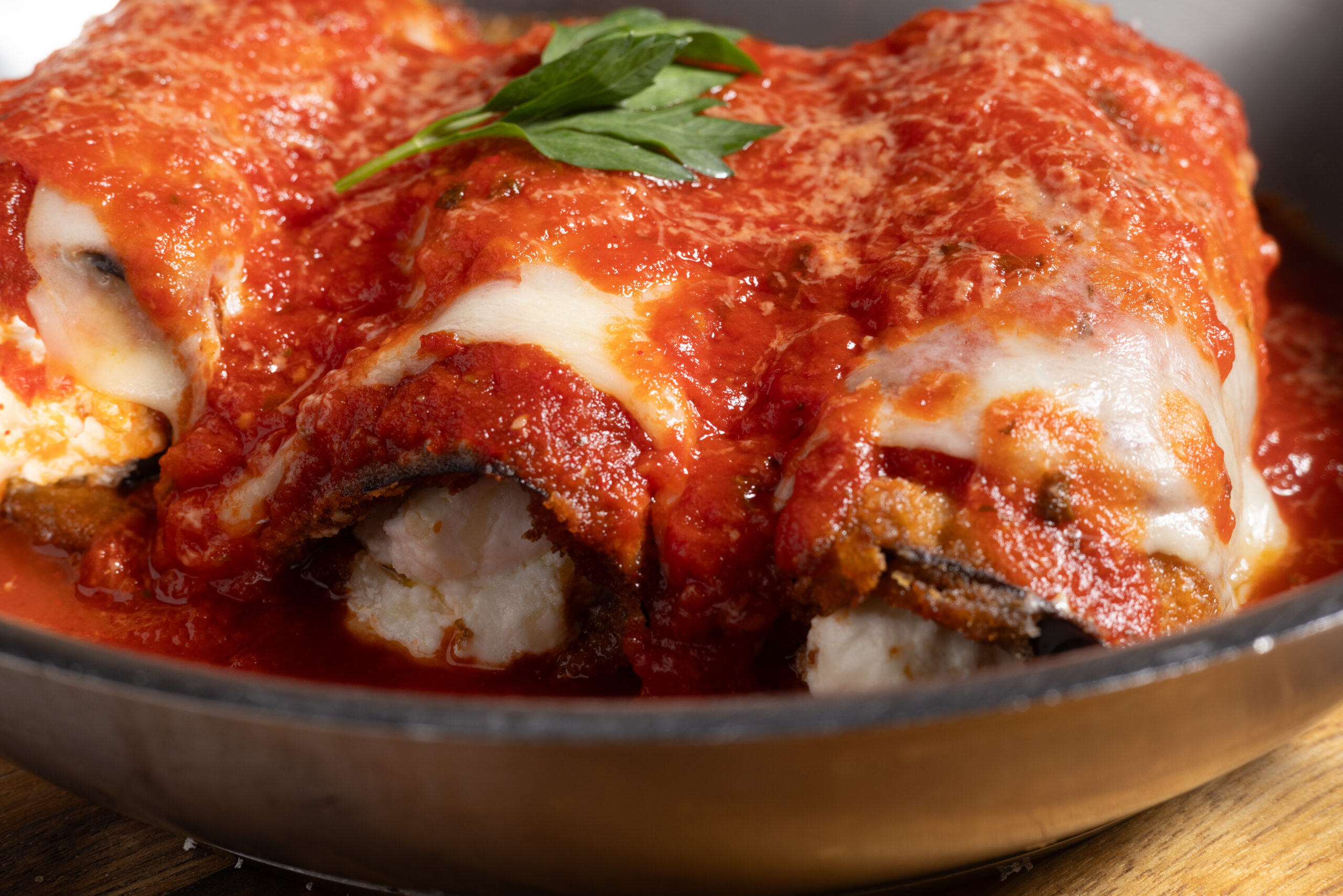 eggplant rollatini – walk-in menu A close-up of baked eggplant rollatini covered in rich tomato sauce and melted cheese, garnished with a sprig of parsley in a pan at TABLE Mercato.
