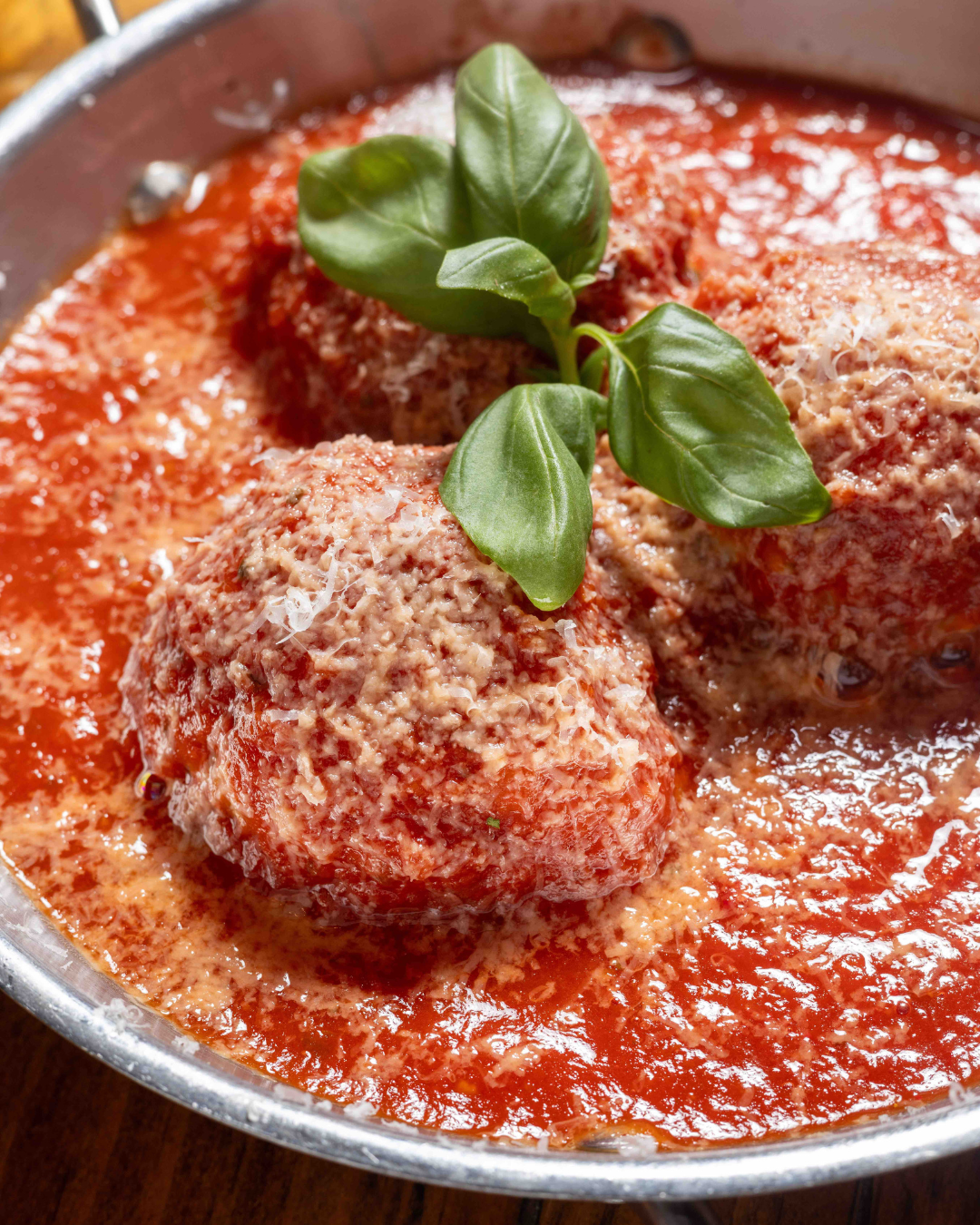 Three meatballs topped with grated cheese in tomato sauce, garnished with basil leaves, served sizzling in a pan. Enjoy this delightful dish after making Reservations at Table Boston for an unforgettable dining experience.