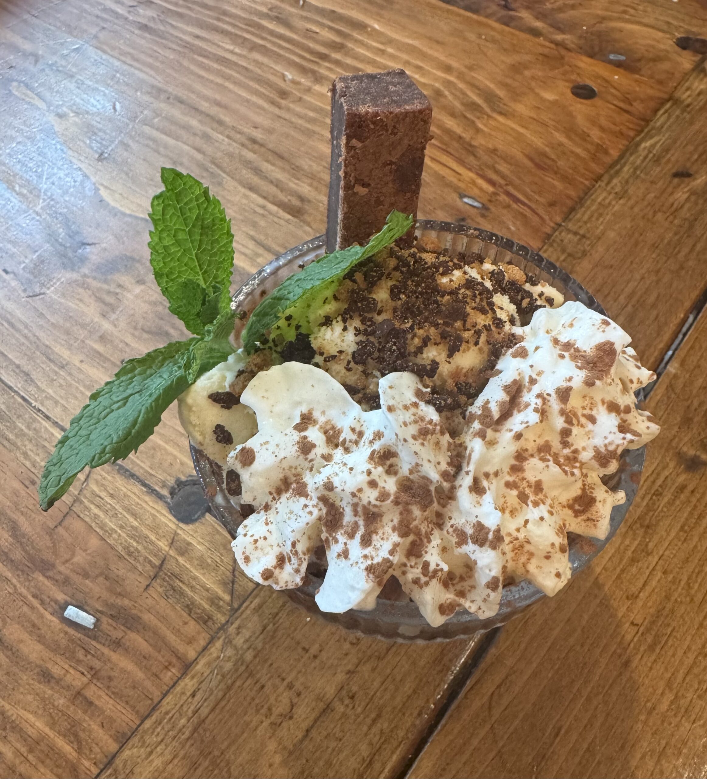IMG_1592 A glass bowl with vanilla ice cream, whipped cream, cocoa powder, a rectangular chocolate piece, banana slices, and a mint sprig on a wooden table—just like you’d find on the Table North End Boston menu.