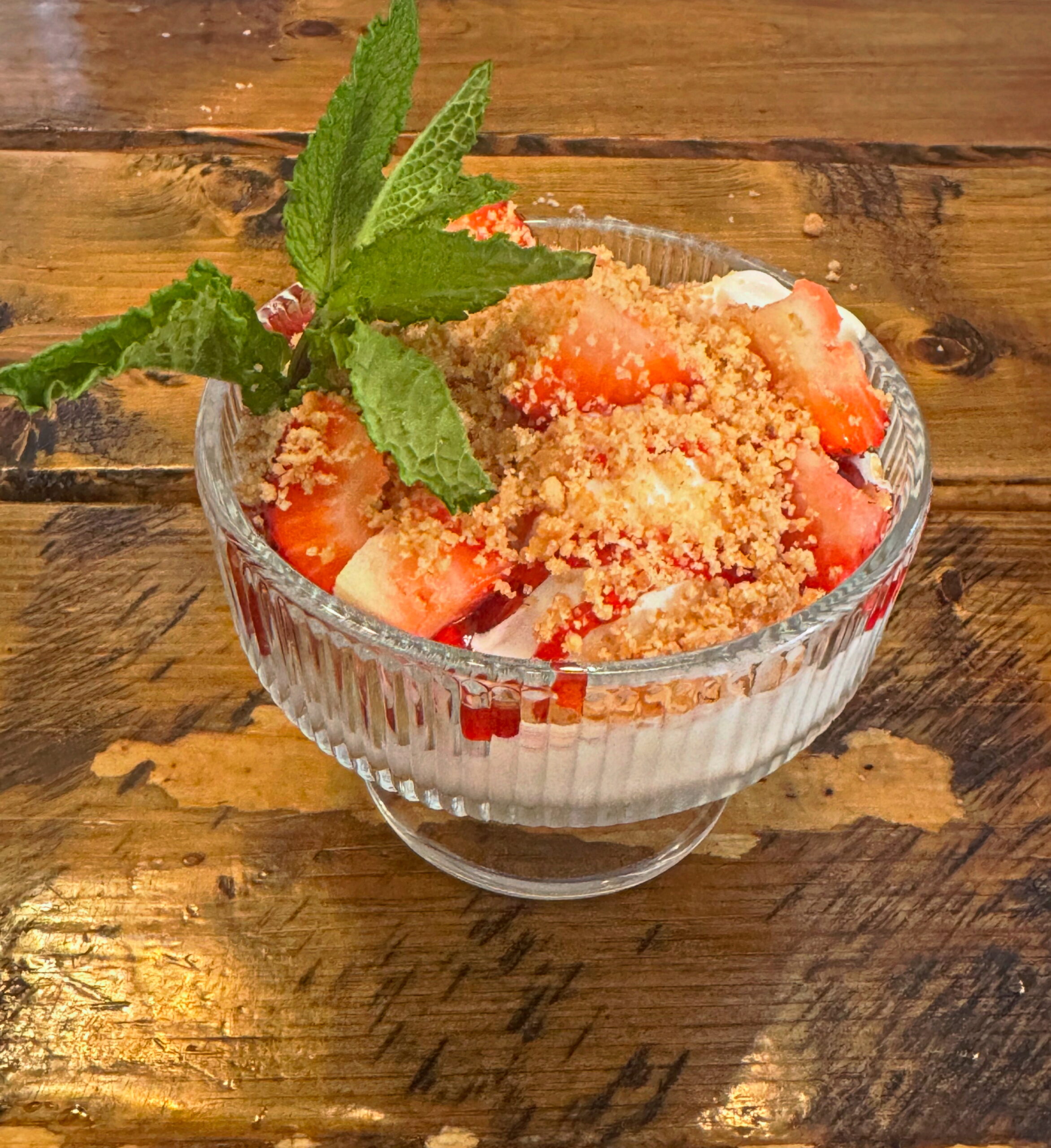 A glass bowl of dessert with whipped cream, chopped strawberries, crumb topping, and a sprig of mint sits on a wooden table—just like the sweet treats featured on the Table North End Boston menu.