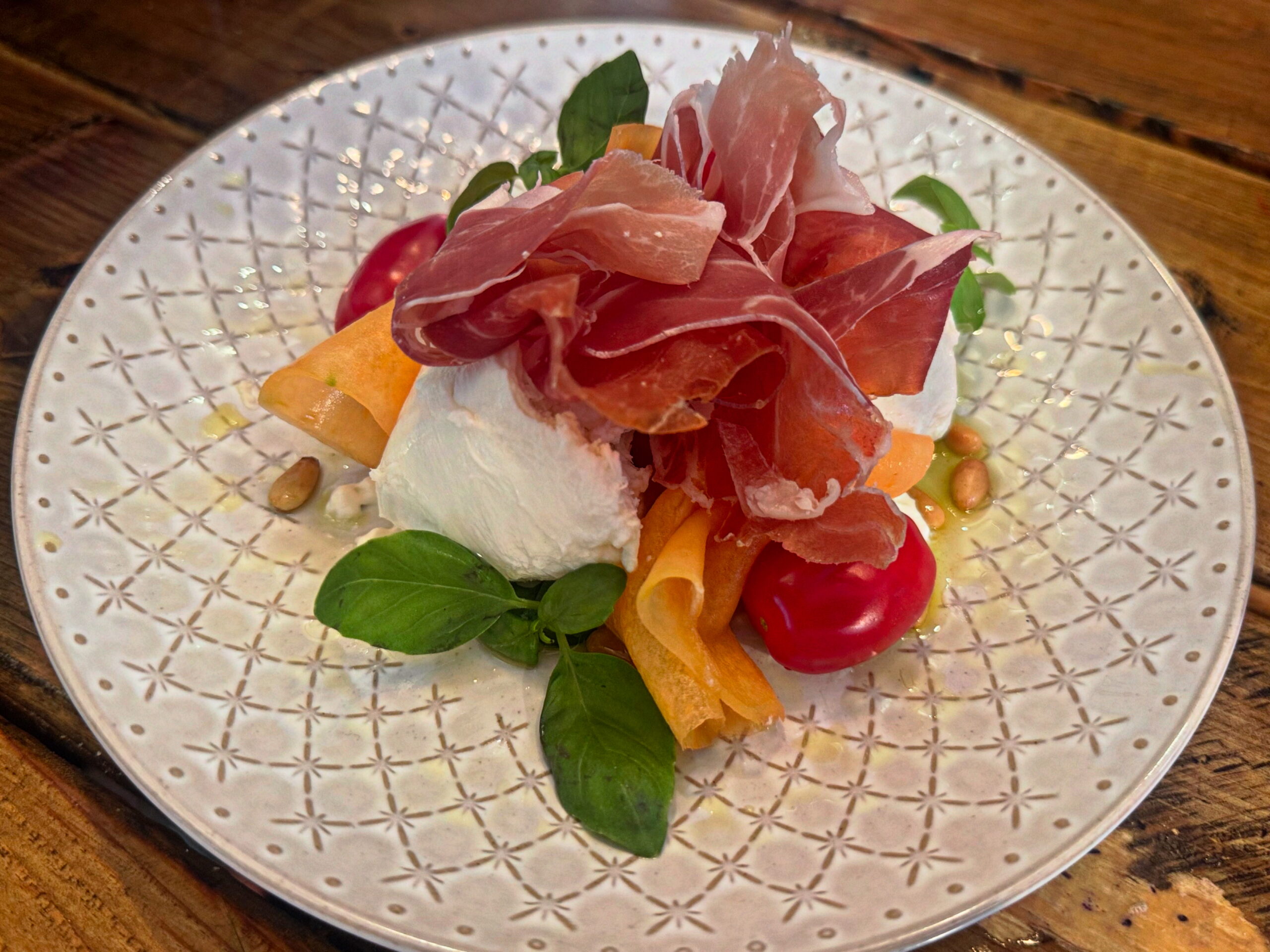 A plate from the Table North End Boston menu featuring prosciutto, mozzarella, cherry tomatoes, carrot ribbons, fresh basil, and pine nuts on a patterned white dish.