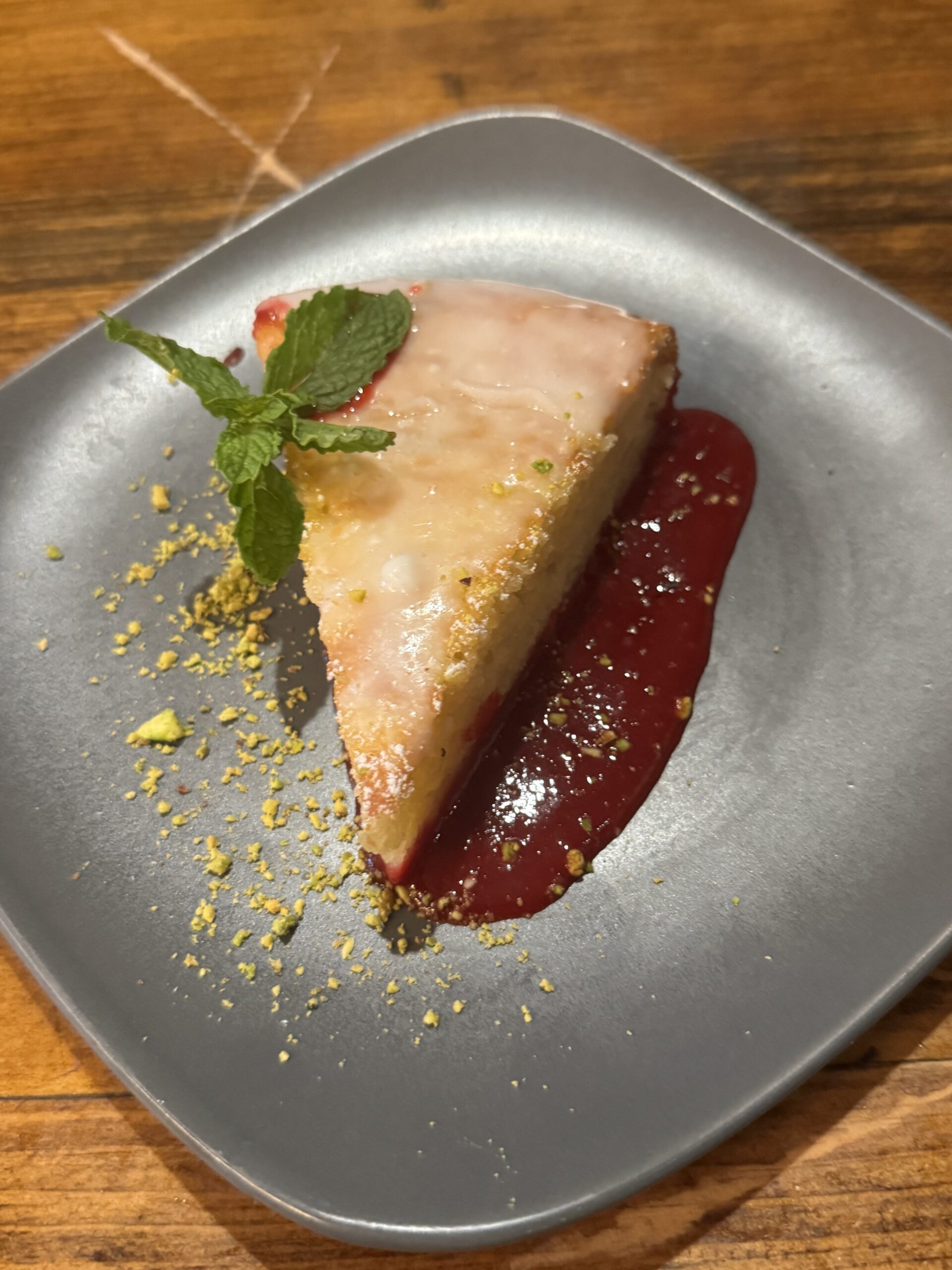 IMG_6918 A slice of glazed cake garnished with a mint sprig, served on a gray plate with berry sauce and sprinkled crushed nuts—just one sweet option from the Table North End Boston menu.