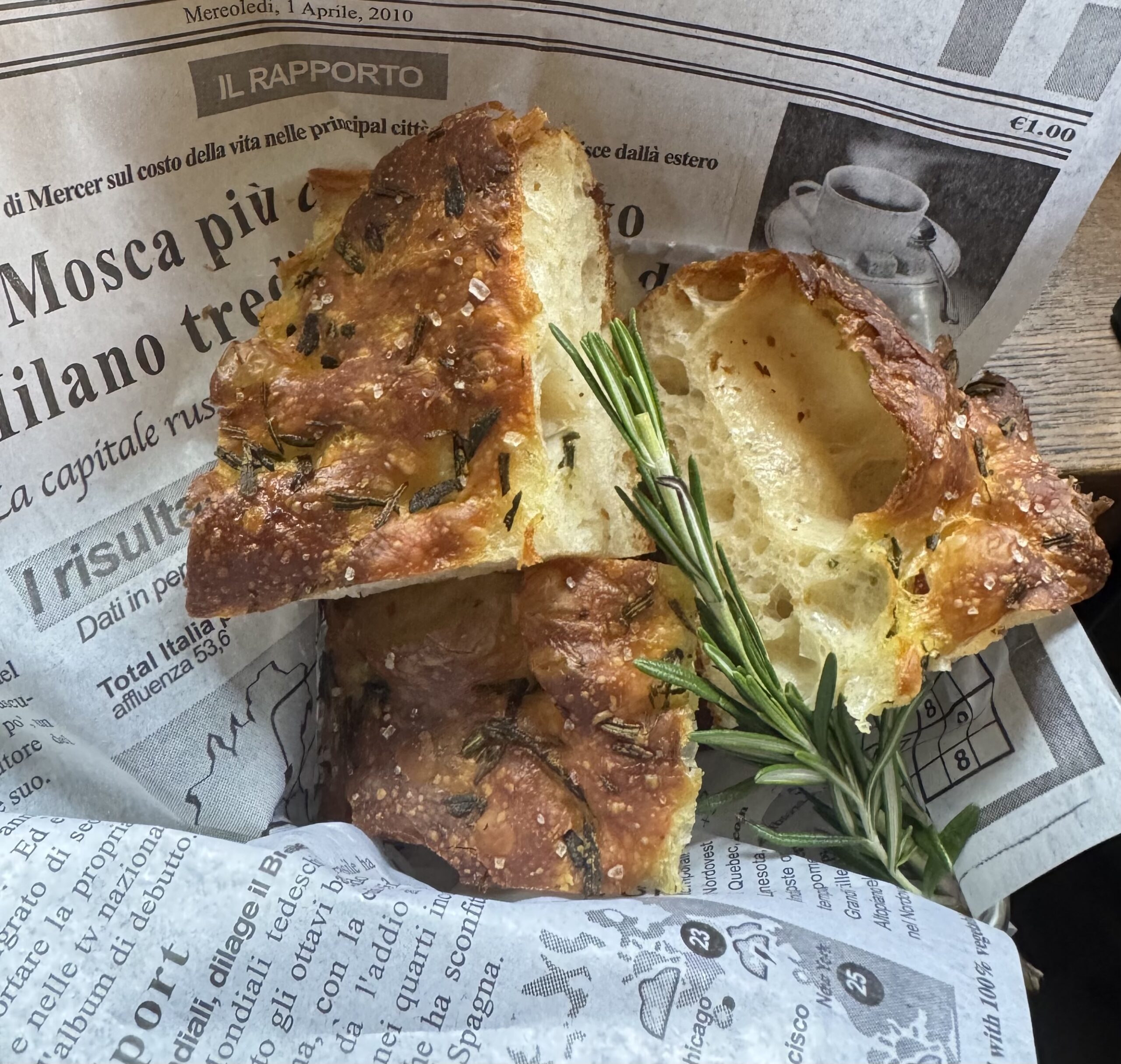 Two pieces of focaccia bread with rosemary rest on a sheet of newspaper, alongside a sprig of fresh rosemary—just like you'll find on the Table North End Boston menu.
